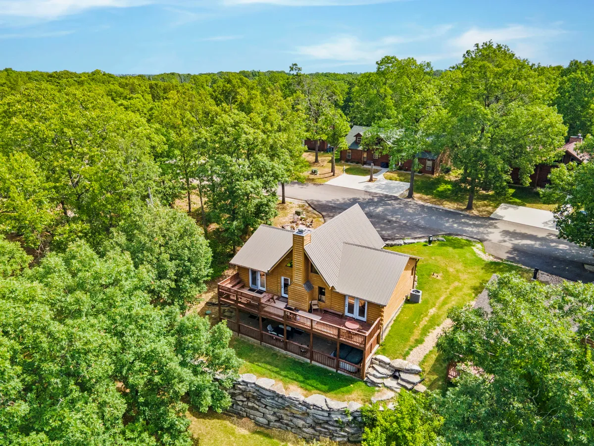 This image features a charming wooden cabin surrounded by lush greenery, with a spacious deck and stone wall visible. The open yard and ample outdoor space suggest plenty of room for outdoor activities, walks, and relaxation in a tranquil natural setting.