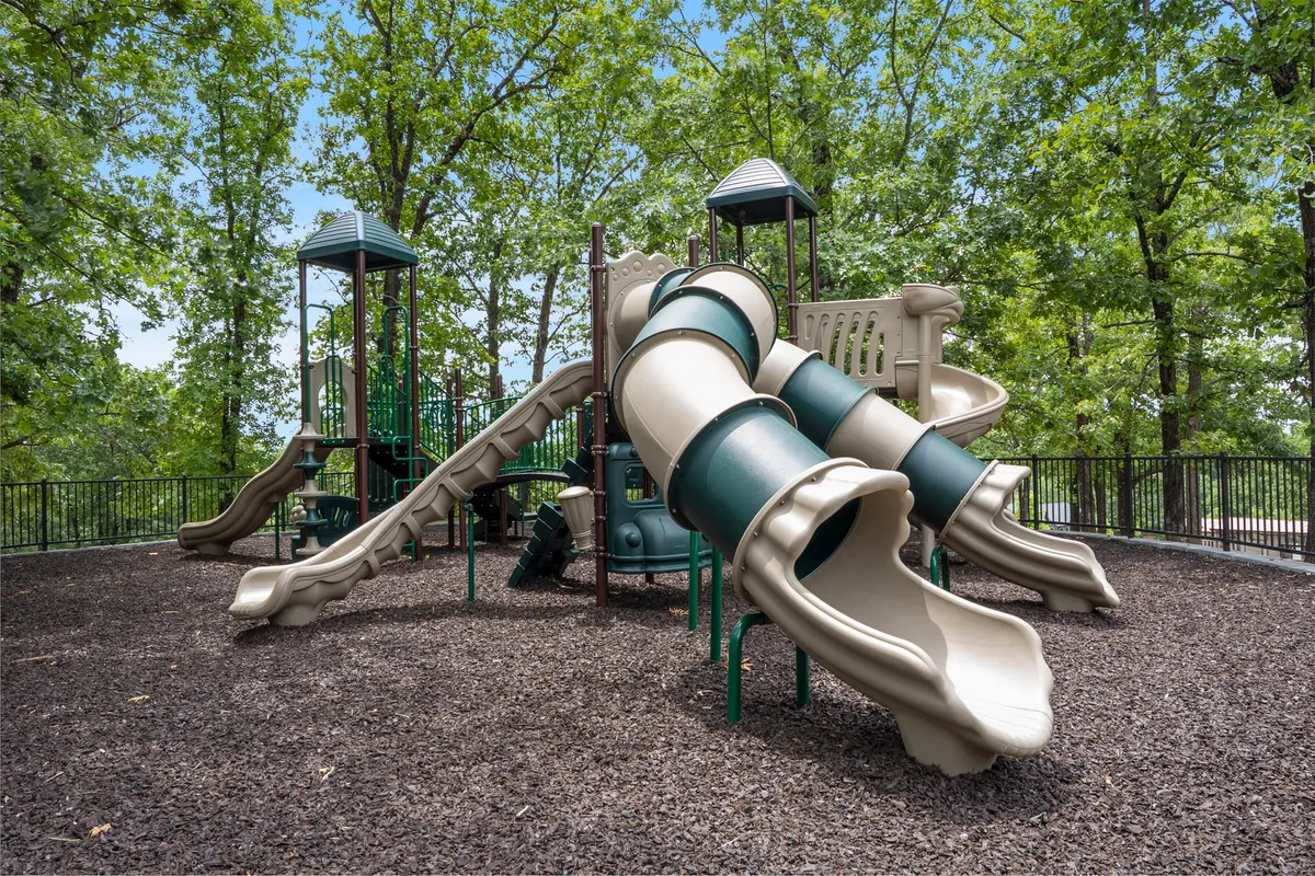 A vibrant playground scene featuring colorful slides, a climbing structure, and mature trees in the background. Children and families are encouraged to play, gather, and picnic, creating a lively, joyful atmosphere.