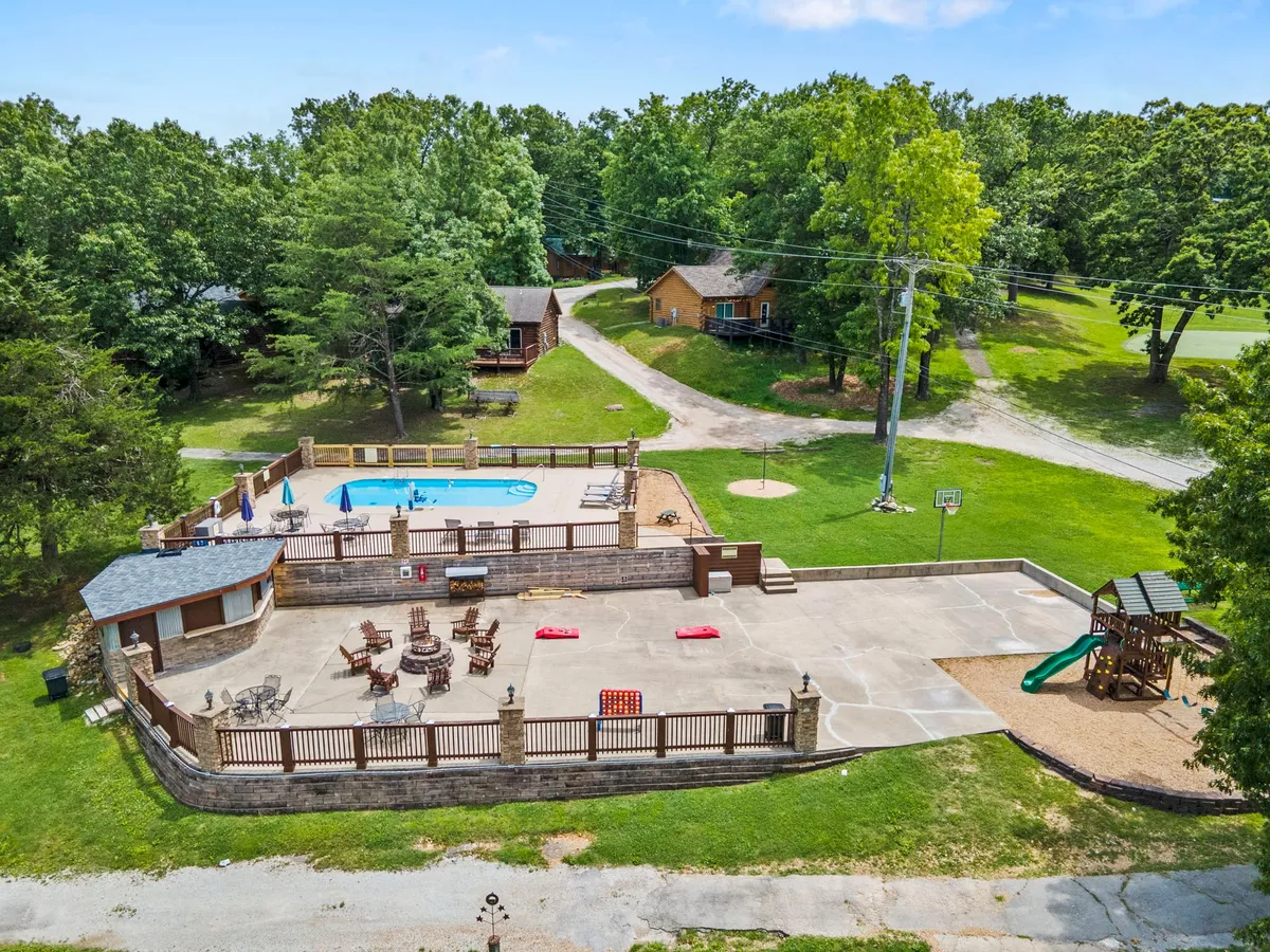 A vibrant outdoor resort scene featuring a swimming pool surrounded by lounge chairs, a playground area, a basketball hoop, and neighboring cabins, all nestled amongst lush green trees. Children play and families relax in the joyful atmosphere.