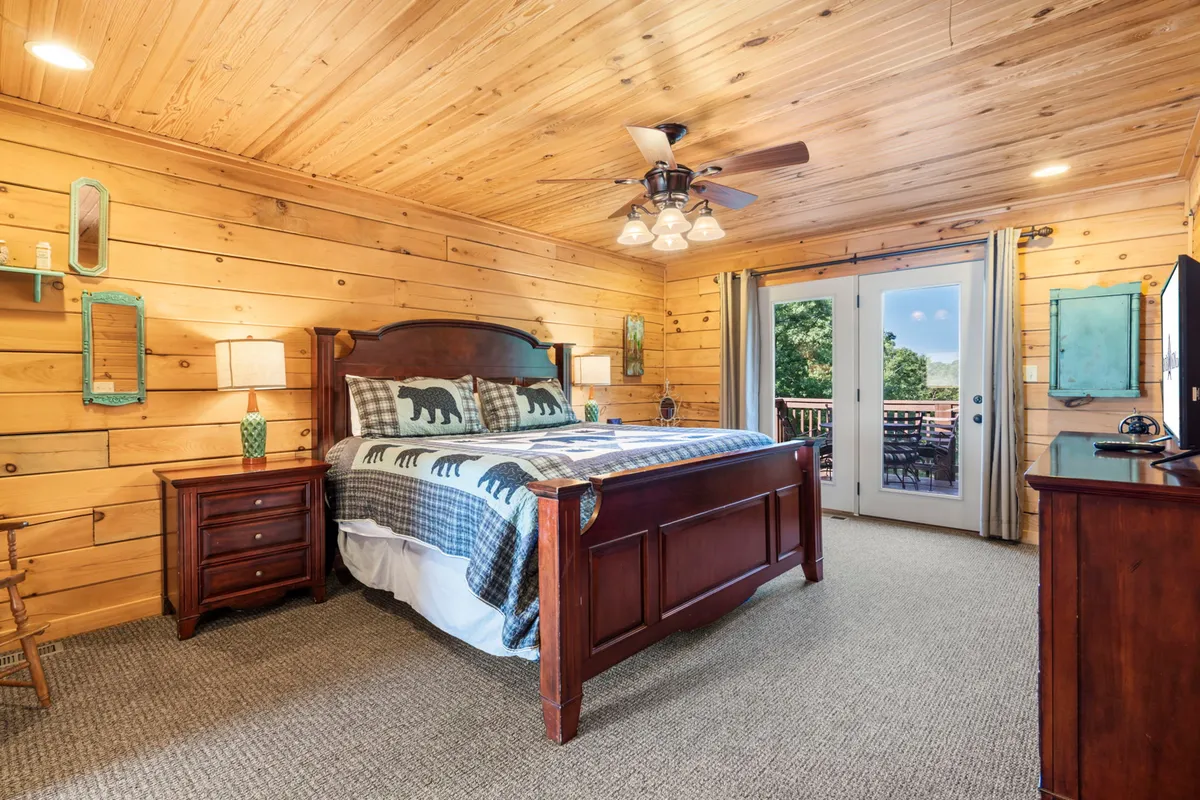 A cozy bedroom with a wooden bed, bear-themed bedding, large windows that let in natural light, and a nightstand with a lamp. The overall ambiance is rustic and comfortable, perfect for unwinding after a day of activities.