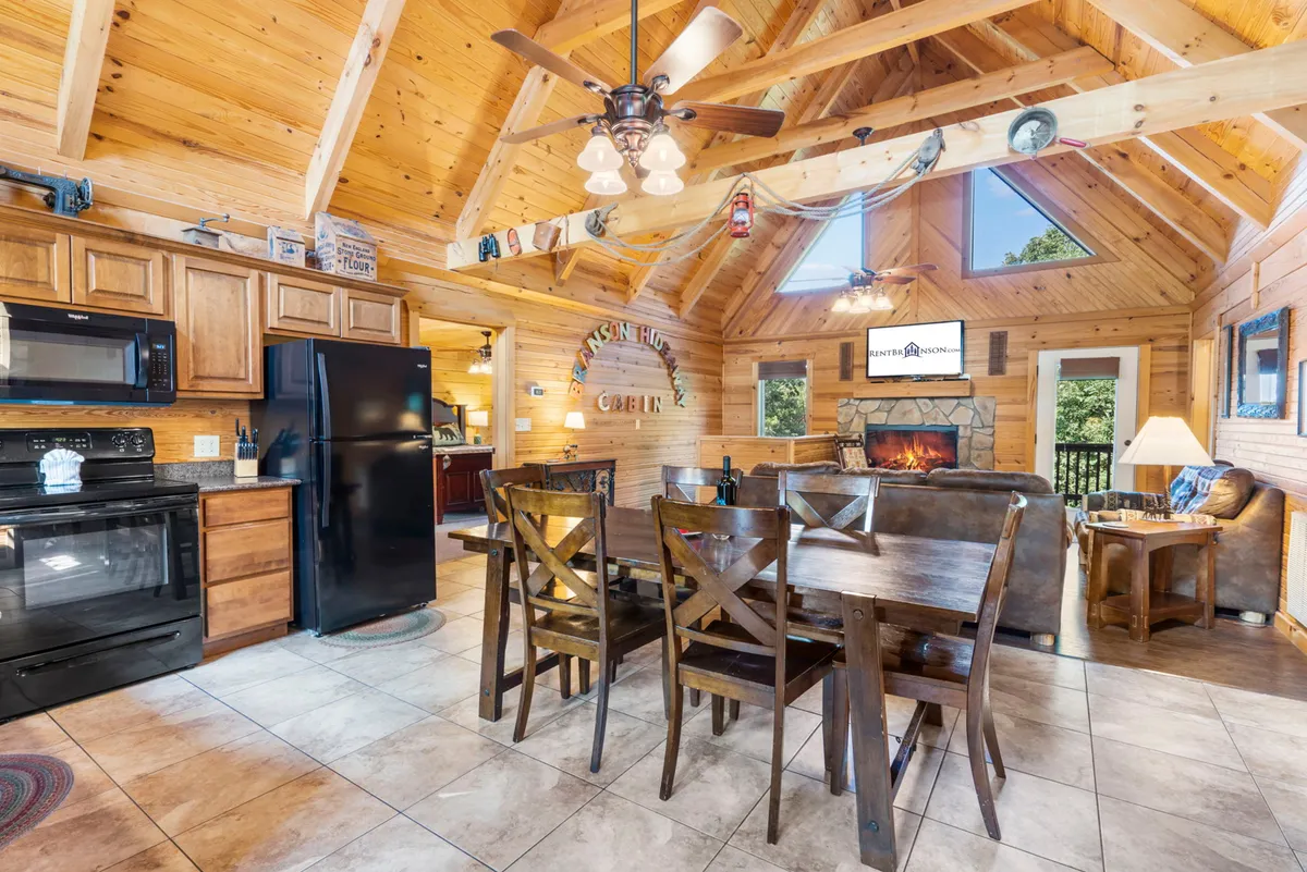 This interior shot highlights a cozy dining area with a wooden table, a stone fireplace, visible kitchen appliances, and large windows bringing in natural views. The space feels welcoming and is set up for both casual family meals and warm gatherings by the fire.