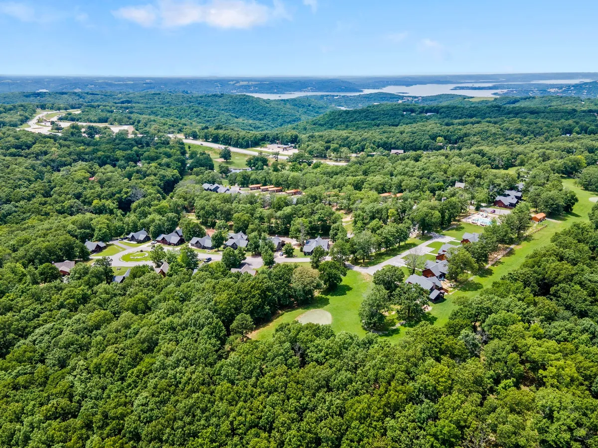 A sweeping aerial view of lush green forests dotted with scattered residential cabins, winding roads, and a distant lake. The landscape is serene and natural, showcasing the untouched beauty of the Ozarks and the proximity to water-based activities.