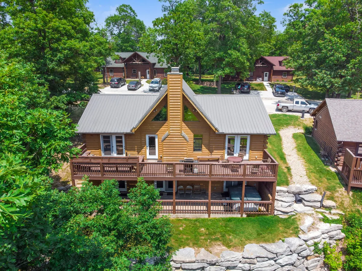 A welcoming log cabin structure with a large wooden deck, surrounded by leafy trees and a stone pathway. The space is set up for outdoor gatherings, barbecues, and relaxation, inviting guests to enjoy the serene outdoor environment.