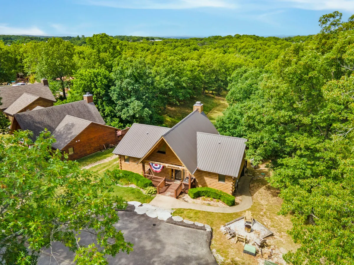 A spacious log cabin nestled among trees, featuring a landscaped pathway leading to a cozy fire pit with seating. The cabin's large deck and inviting porch suggest ample space for gatherings, outdoor meals, and celebrations.