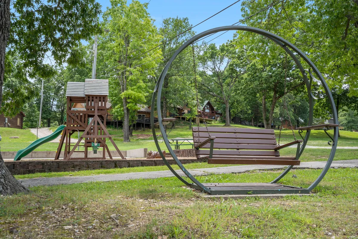 A playful outdoor scene featuring a circular swing and playground structure nestled among green trees, with log cabins visible in the background. The area is relaxed and shaded, ideal for family and pet recreation.