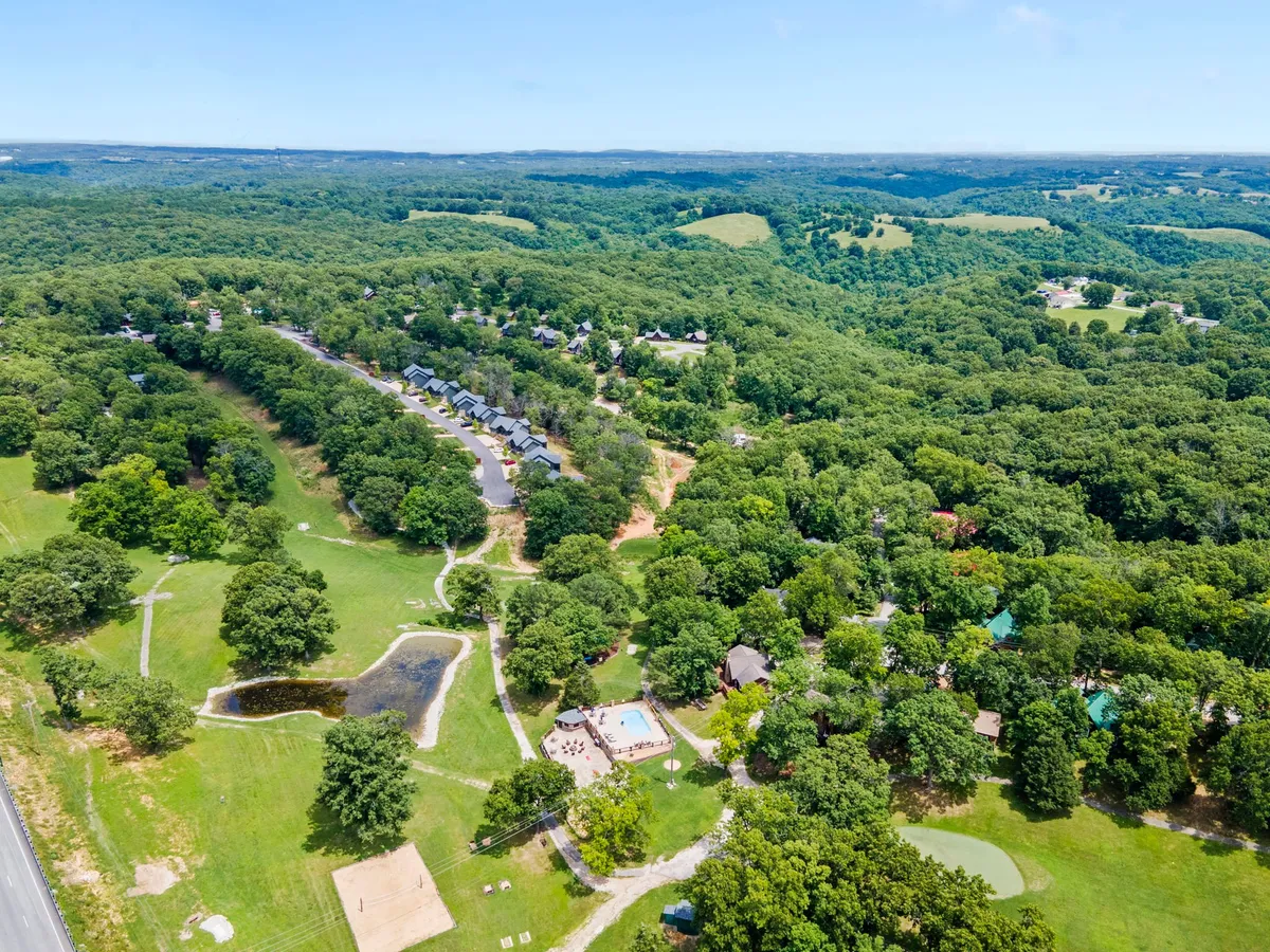 A sweeping aerial view showcasing lush green hills dotted with neatly arranged cabins, a sparkling swimming pool, a serene small pond, and visible outdoor activity areas. The communal nature of the resort is highlighted by the open spaces and inviting amenities situated amidst a scenic Ozarks landscape.
